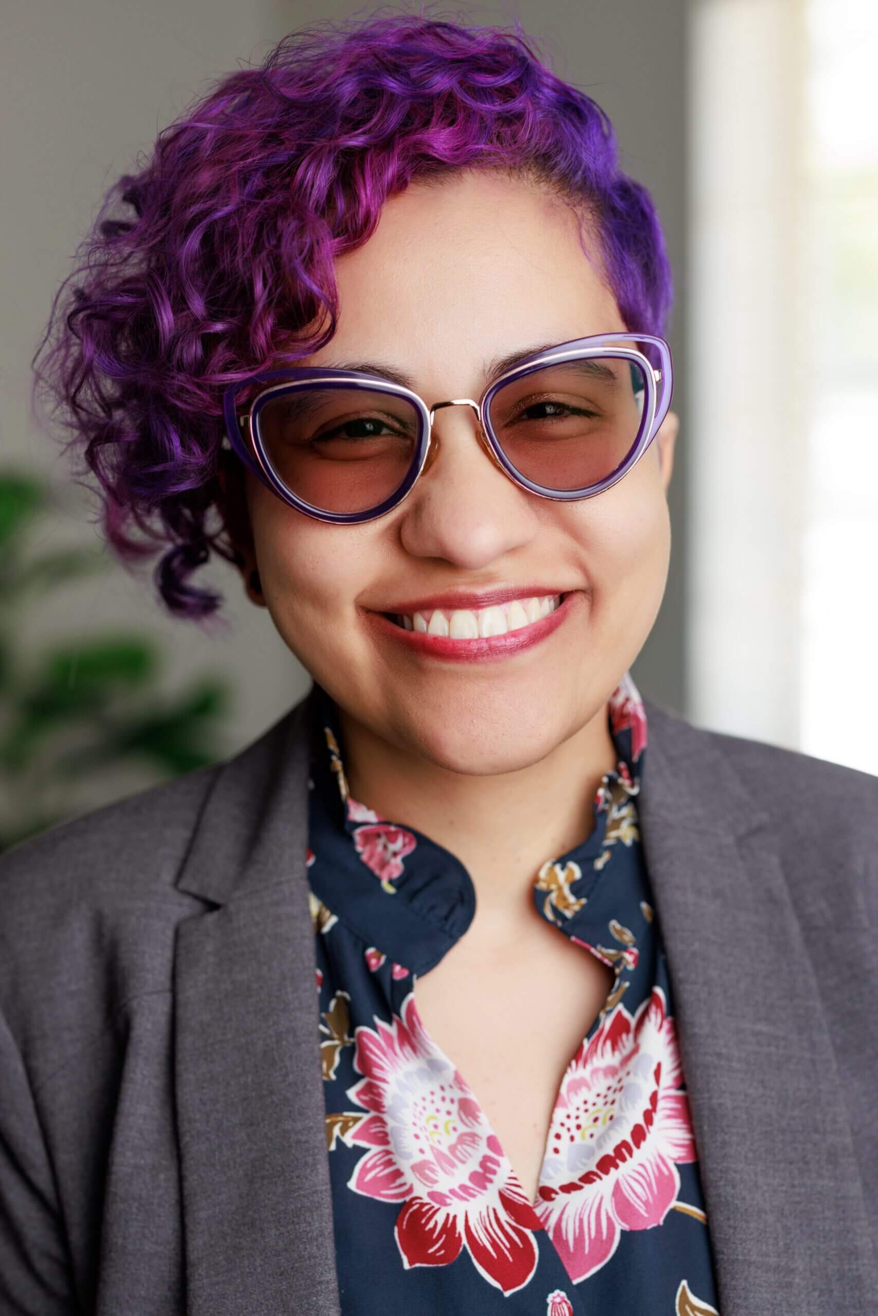 Person with purple hair wearing sunglasses, smiling.